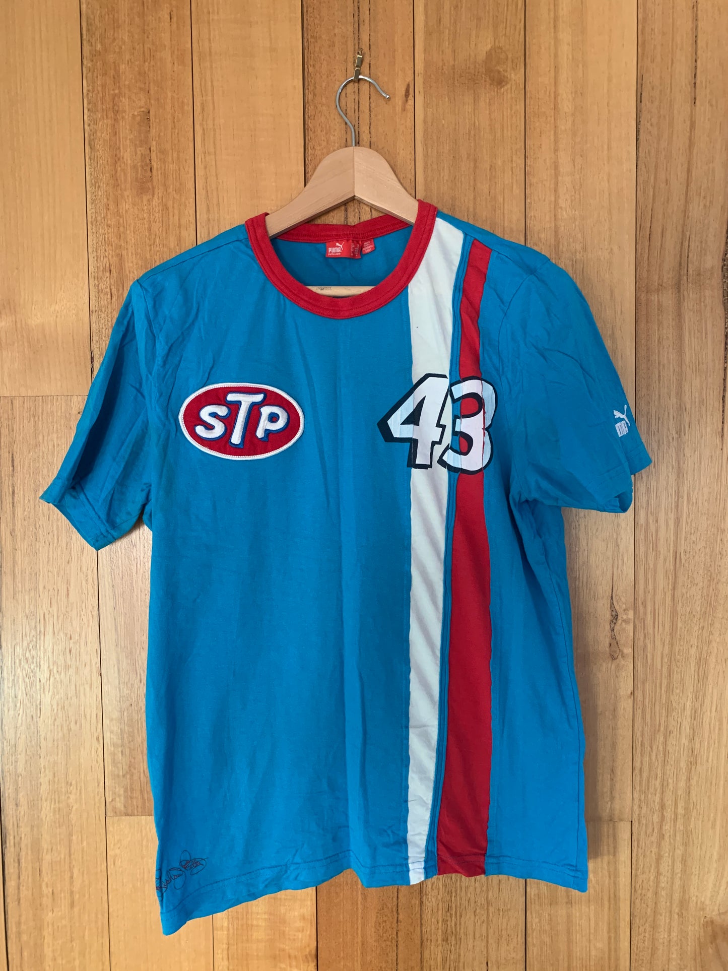 A blue Puma Richard Petty STP Racing T-Shirt with the number 43 and the STP logo on it, displayed on a wooden hanger.
